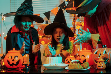 Asian family mom, dad in a mask, and daughter celebrate Halloween together, sharing a cake and carved pumpkins under atmospheric, warm-toned festive lighting and witch hats.