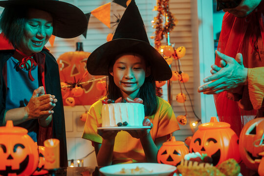 Asian family mom, dad in a mask, and daughter celebrate Halloween together, sharing a cake and carved pumpkins under atmospheric, warm-toned festive lighting and witch hats.