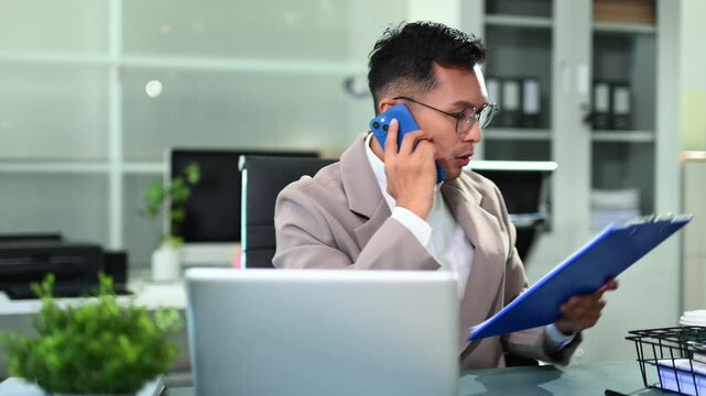 Businessman managing multiple tasks, talking on phone, checking reports, and working on laptop in a busy modern corporate office.