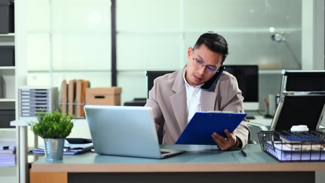 Businessman managing multiple tasks, talking on phone, checking reports, and working on laptop in a busy modern corporate office.