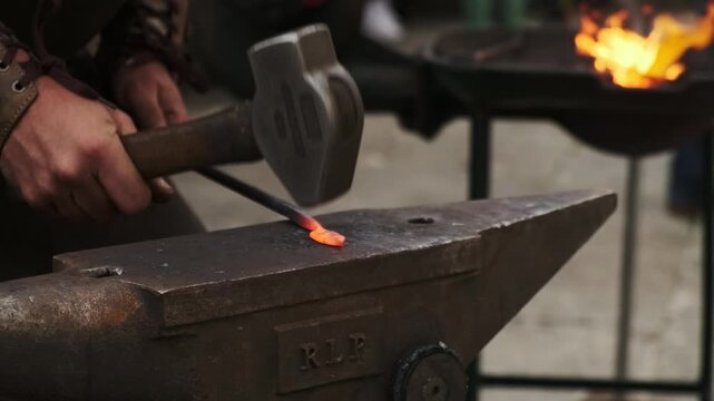 Static close-up of a blacksmith striking red-hot iron on an anvil with a hammer; the coal forge blazes softly out of focus in the background.