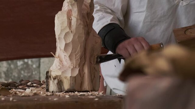 Static shot with a blurred object in the foreground as an artisan carves a log with chisel and mallet in the background; tools and wood shavings scattered across the table.