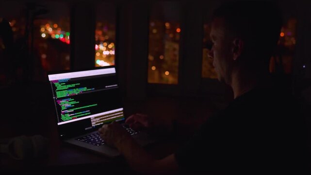 A young male programmer in front of a window with a lights of night city, on his laptop screen is code. out of focus. 