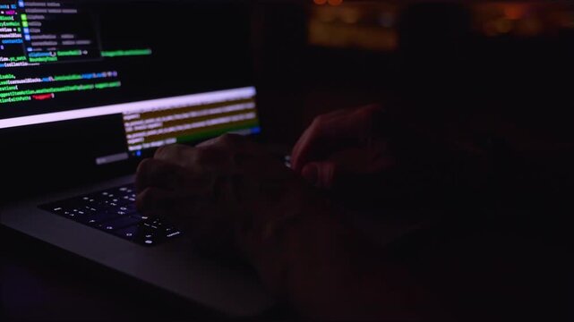 A young male programmer in front of a window with a lights of night city, on his laptop screen is code. out of focus. 