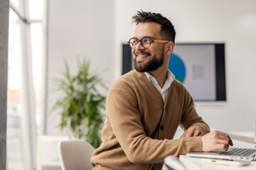 Obraz premium Bearded Professional In Brown Cardigan Working At Modern Office Desk With Natural Light