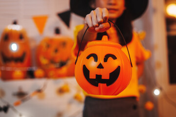 A cheerful young girl in a witch hat holds a pumpkin bucket toward the camera, ready for trick-or-treating amidst vibrant, spooky Halloween decorations and glowing jack-o'-lanterns.
