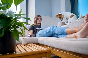 Woman lying on terrace sofa using laptop with mouse and coffee cup on wooden table. Female freelancer working remotely on patio with her dog nearby