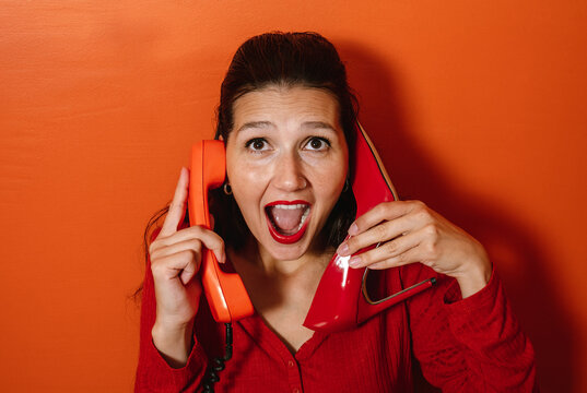 Woman holding phone and high heel looking surprised against orange background