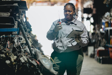 Female technician check used car damaged engine block at scrapyard warehouse recycle area part. African American engineer inspecting rust oily auto motor old spare part in junkyard for reuse service