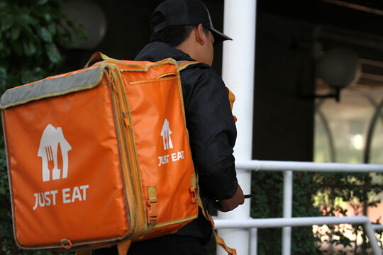 Just Eat pedir comida a domicilio con repartidor de plataforma entrando en portal de la direcci&oacute;n de cliente para reparto con bolsa naranja de entrega y pedido app m&oacute;vil de restaurante marketplace  