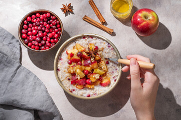 Hand holding spoon in to plate of oatmeal with apple, cranberry and cinnamon on a light background