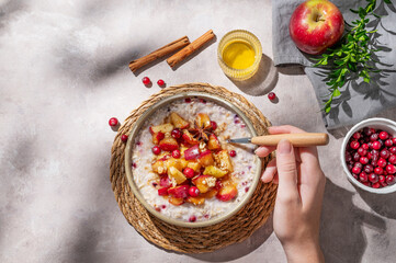 Hand holding spoon in to plate of oatmeal with apple, cranberry and cinnamon on a light background