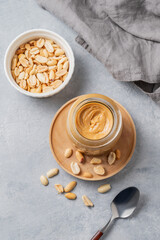 Creamy peanut butter in a jar on a wooden plate on a blue background with a scattering of roasted nuts