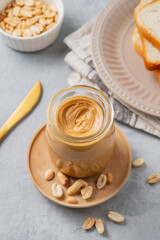 Creamy peanut butter in a jar on a wooden plate on a blue background close up with a scattering of roasted nuts
