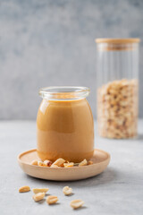Creamy peanut butter in a jar on a wooden plate on a blue background with a scattering of roasted nuts.