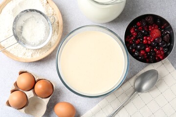 Liquid dough in bowl, berries and ingredients on light grey table, flat lay