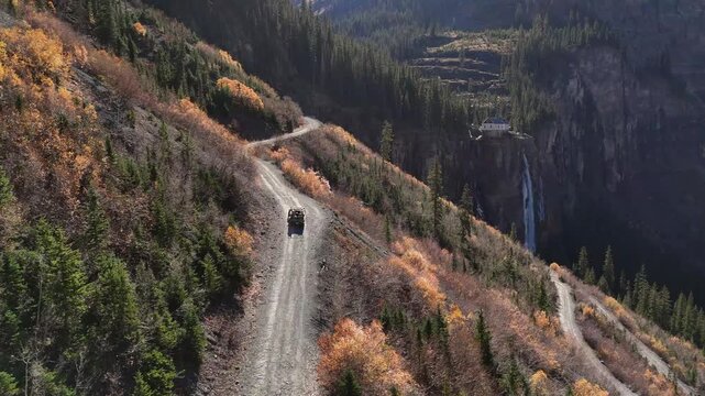Aerial view of a vehicle navigating Black Bear Pass, a winding dirt road amidst the rugged terrain, with Bridal Veil Falls cascading in the background, Telluride, Colorado, United States.