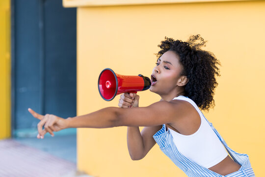 Young woman shouting through red megaphone and gesturing in front of yellow wall
