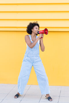 Woman with curly afro hair shouting through megaphone in front of yellow wall