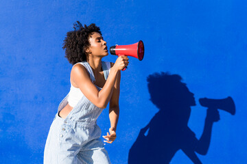 Woman with curly afro shouting into red megaphone against blue wall