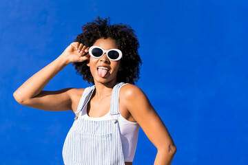Playful woman with afro and sunglasses posing joyfully against blue wall