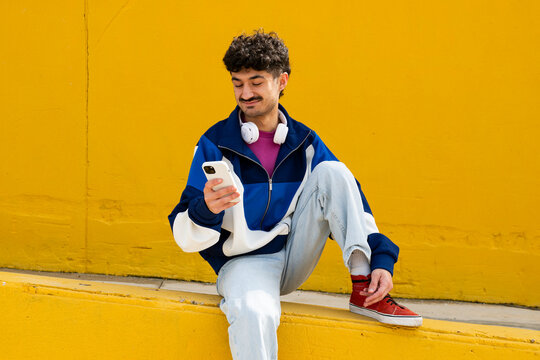 Young man in urban clothing using smartphone with wireless headphones against a yellow background