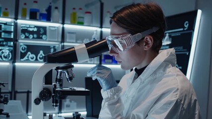 Young scientist in a clean suit analyzing a sample through a microscope. Futuristic laboratory with advanced equipment and digital displays