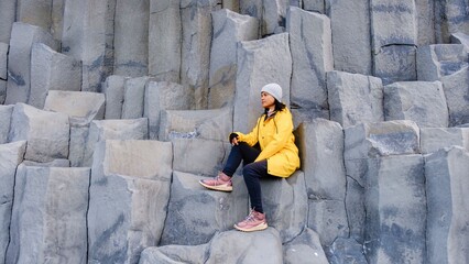 An Asian woman in a bright yellow jacket rests on dramatic basalt columns at Reynisfjara Beach in Iceland. The striking rock formations create a mesmerizing backdrop against the rugged coastline.
