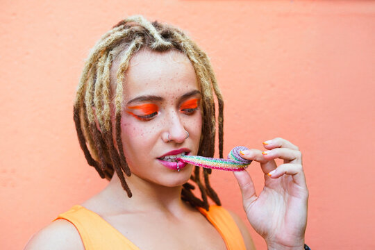 Smiling woman with dreadlocks eating rainbow candy with bold orange makeup