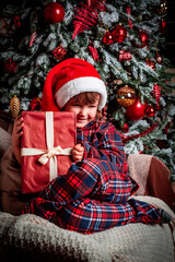 Happy little girl child in pajamas and festive santa hat smiling while sitting near christmas tree with gift box. Magic. New Year's eve. St. Nicholas Day. Present. Time of holidays and miracles.	