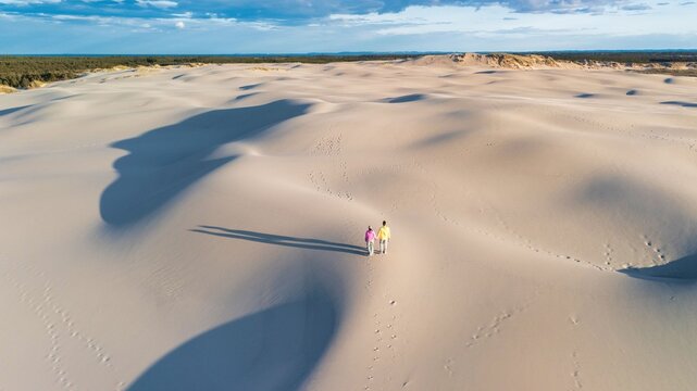 A couple stroll hand in hand across the expansive, undulating white sand dunes of Denmark. The warm golden light of sunset casts long shadows, Rabjerg Mile Dunes, Denmark.