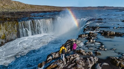 Two adventurers stand on rocky terrain near Dettifoss, observing the majestic Selfoss waterfall surrounded by a rainbow. The vibrant colors contrast beautifully with the rugged landscape of Iceland