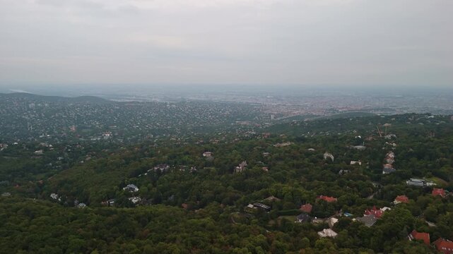 Forward ascending drone shot from Normafa towards Budapest, Hungary, revealing the city skyline and Danube River over the forested Buda Hills under cloudy skies.