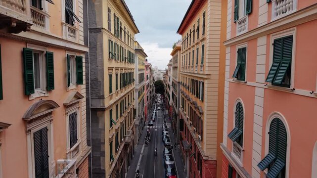 A drone flies between tall colorful buildings along a narrow street in Genoa, Italy, showing traffic and architecture with shuttered windows and warm facades.