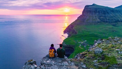 A couple watching sunset Gasadalur waterfall as it tumbles down rocky cliffs into the Atlantic...