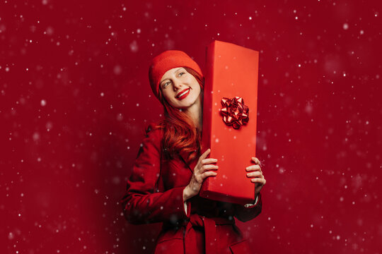 Joyful woman holding Christmas gift box in red winter outfit and snow