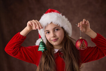 Girl hold in hands Christmas tree balls decoration and dressed in Santa Claus hat, holiday portrait in red background
