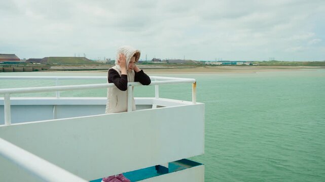 Girl standing alone on upper ferry deck during windy day in Dunkirk port France. Child protecting head with hood while surrounded by sea and coastline. Young traveler enjoying breezy boat ride near