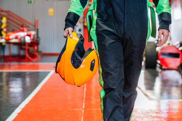 Racer wearing a suit holding an orange helmet, walking on the red floor of a go kart pit lane or garage