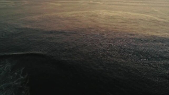 Aerial view of the dark ocean water reflecting the warm tones of the sunset as it meets the horizon creating a gradient of colors, Hossegor, France.
