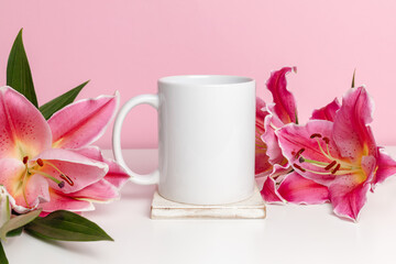 White mockup mug with gentle pink lily flowers on pink background. Mockup ceramic cup with copy space for cute design or print
