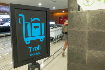 A bold blue sign shows a luggage trolley icon in an airport baggage area, guiding travelers to trolleys.