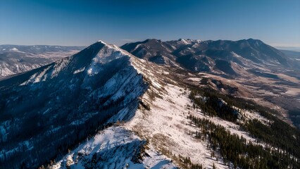 Snowy mountain ridge seen from above, with forested slopes, open valleys and distant ranges under a clear blue sky. Concept Aerial view of snowy ridge, Forested slopes and valleys