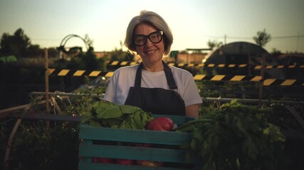 Happy senior female farmer holding a wooden crate full of fresh organic vegetables, smiling proudly in her garden at sunset, embodying a healthy and sustainable farm to table lifestyle