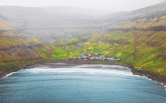 Aerial view of a quaint village nestled by the dark blue sea, embraced by vibrant green hills under a misty sky, Tj&Atilde;&cedil;rnuv&Atilde;&shy;k, Streymoy, Faroe Islands.