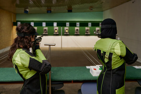 Two women practicing shooting at an indoor range, aiming rifles at targets, competitive sport, precision and focus, professional training for shooting competition, indoor activity.