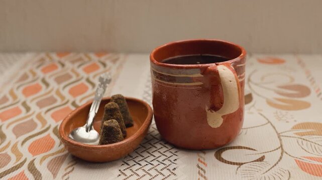 Clay mug with black coffee and a small dish holding brown sugar (piloncillo) and spoon on a patterned cloth