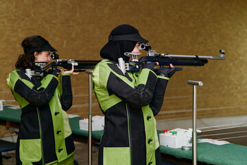 Two female athletes aiming air rifles in shooting competition, demonstrating focus and precision in indoor sports, showcasing skill and determination in target practice, indoor setting.