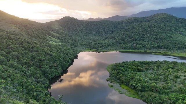 Cinematic aerial shot at sunset over Rio Capricornio near Caraguatatuba, Brazil. Smooth pan with river reflections, clouds, and golden light creating a tranquil tropical scene.