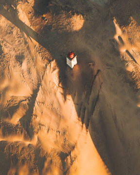 Aerial view of a stark white lighthouse with a red roof standing defiant against the shifting sands of Rubjerg Knude, a beacon amidst nature's relentless embrace, L&Atilde;&cedil;kken, Denmark.